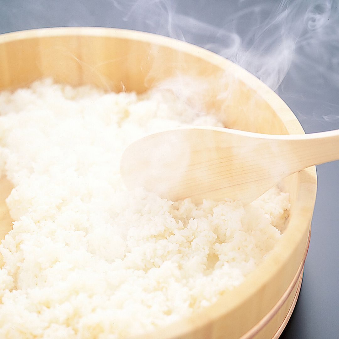 Steaming white rice in a light natural wood bamboo steamer basket with a wooden spoon resting inside, set against a dark gray background.