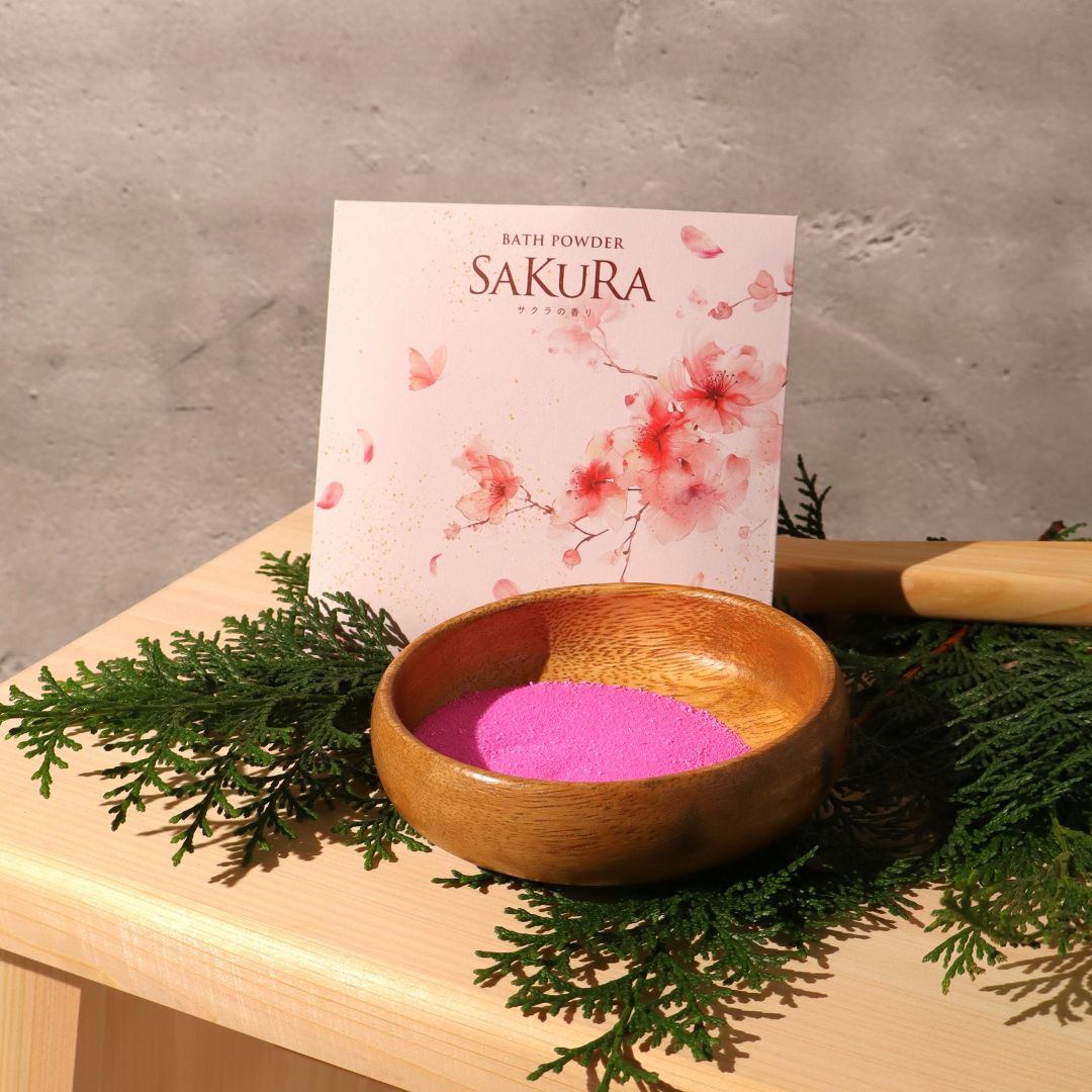 Bowl of pink bath powder with Sakura packaging on a wooden surface with greenery.