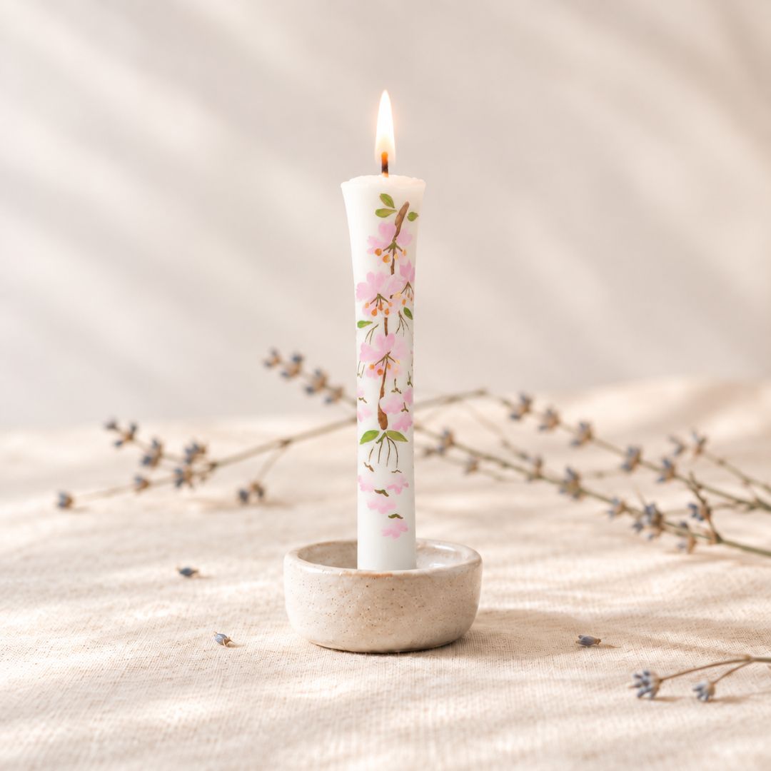 A lit white taper candle with a pink cherry blossom floral print, standing in a small textured ceramic holder on a beige fabric surface with dried branches in the background.