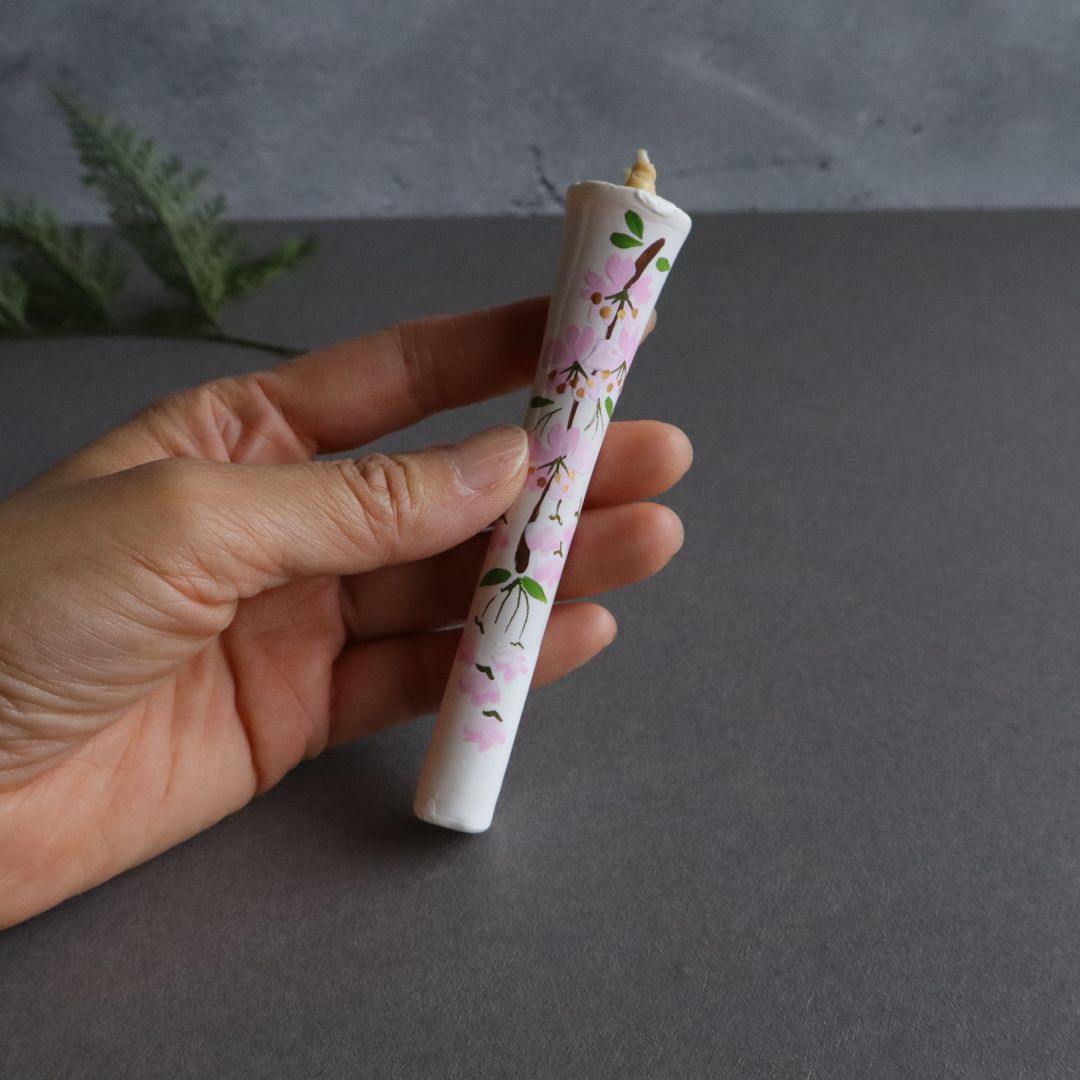 A hand holding a Japanese cherry blossom hand-painted taper candle, with a green fern in the blurred background on a grey surface, symbolizing organic and artisanal home decor.