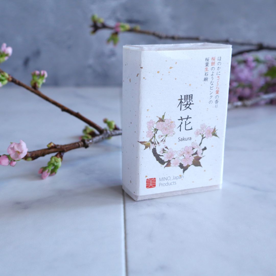 Elegant packaging of handmade Sakura Leaf Soap, featuring delicate cherry blossom illustrations and Japanese calligraphy, displayed on a marble surface with blooming sakura branches in the background.
