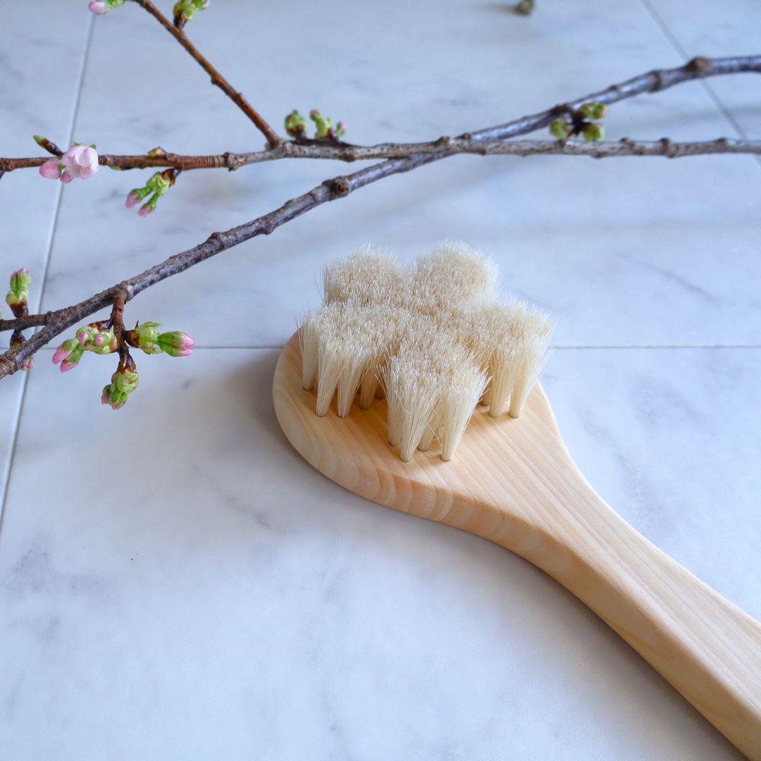 Natural Japanese body brush with soft white horsehair bristles shaped like a sakura flower, set on a light wooden handle, displayed on a marble surface with blooming cherry blossom branches in the background.