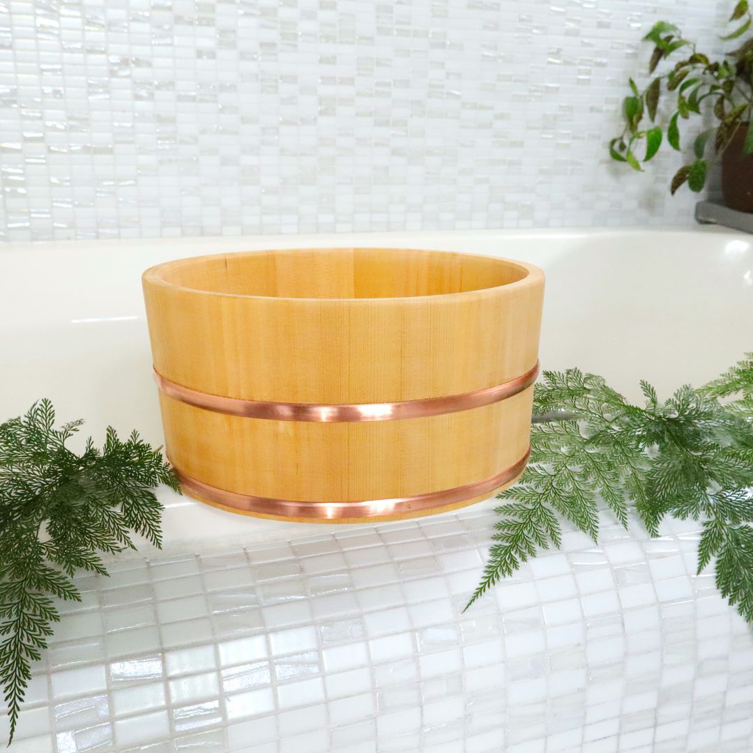 A φ22cm / 8.66" handy bath bucket made of Sawara Japanese cypress sits on a white tiled surface with green ferns around, showcasing the bucket's smooth grain and copper bands.