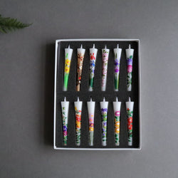 A square photo of a collection of twelve hand-painted sumac wax taper candles, each with a unique organic floral design, arranged in two rows within a white box with black sponge lining. The candles are displayed on a dark gray background with the corner of a green fern leaf subtly intruding into the frame at the top left, emphasizing the natural theme. Each candle measures 10cm or 4 inches in length, presenting a colorful and artisanal appearance.