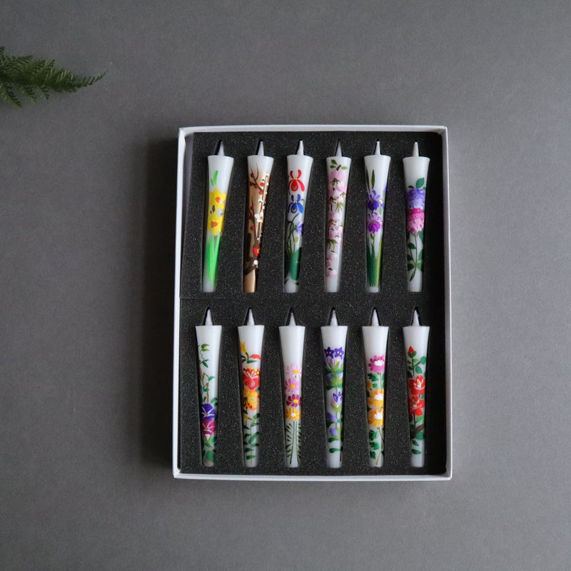 A square photo of a collection of twelve hand-painted sumac wax taper candles, each with a unique organic floral design, arranged in two rows within a white box with black sponge lining. The candles are displayed on a dark gray background with the corner of a green fern leaf subtly intruding into the frame at the top left, emphasizing the natural theme. Each candle measures 10cm or 4 inches in length, presenting a colorful and artisanal appearance.