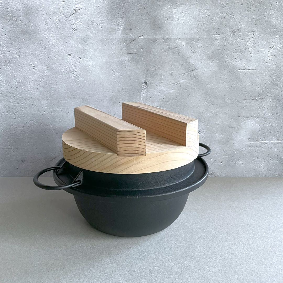 In a grey room, a rice pot made of black cast iron with a wooden lid on top