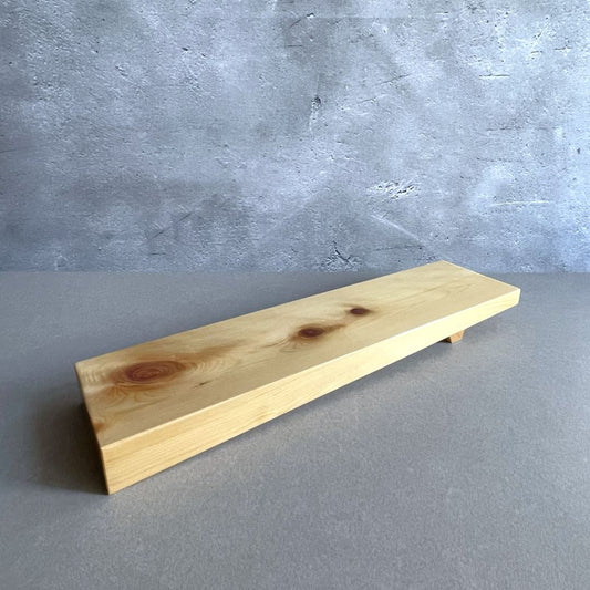 A simple wooden shelf with visible grain and knots is mounted against a textured concrete wall. The shelf is placed at an angle, providing a view of its thickness and the support at the bottom. The scene has a minimalist aesthetic with a focus on natural materials.