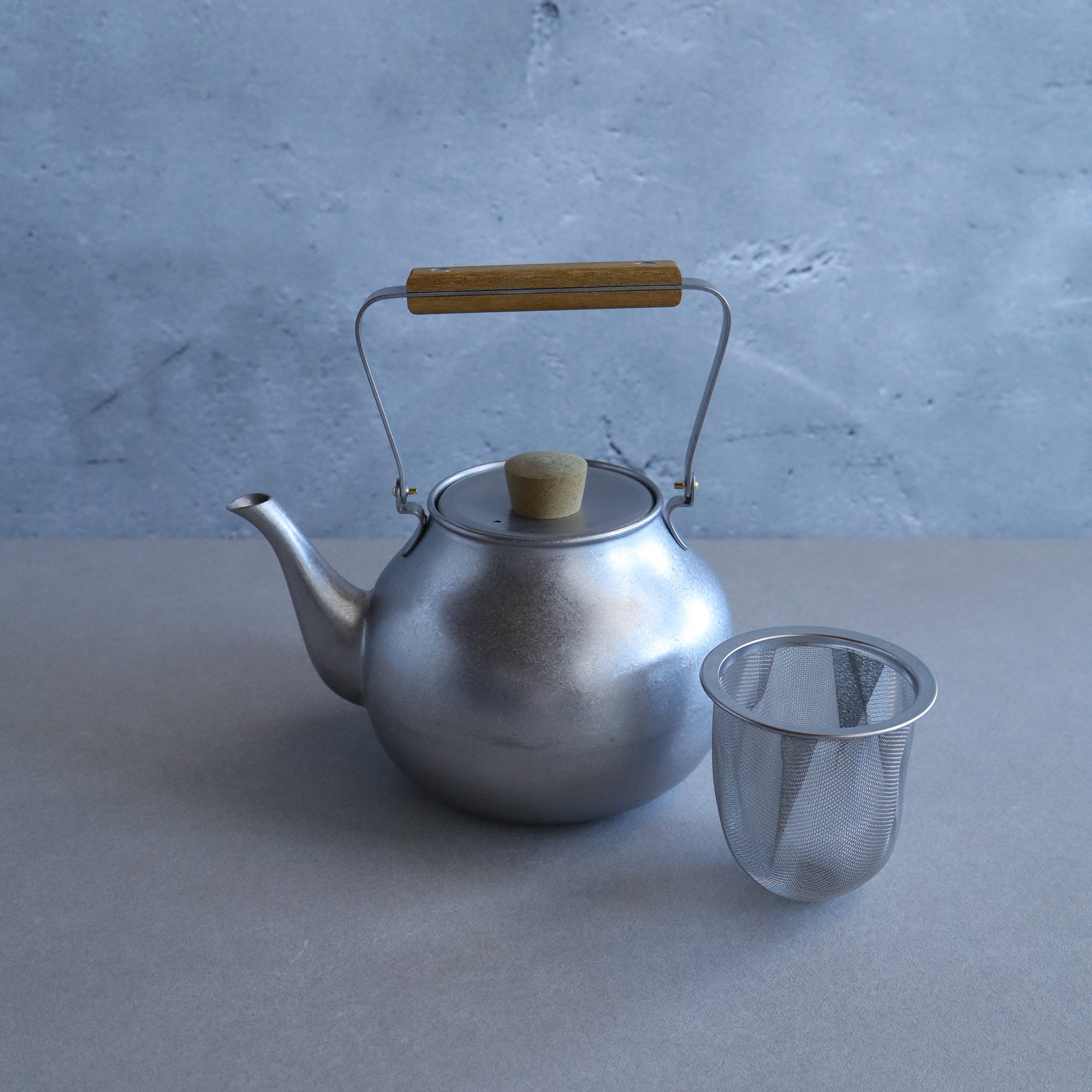 Silver teapot with wooden handle and metal mesh strainer on a gray surface.