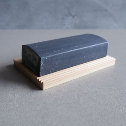 A black charcoal facial soap bar with a smooth, rectangular shape resting on a light-colored hinoki wood soap dish with a grooved design. The soap has a slightly rough texture on the edges, and the background is a neutral gray surface.