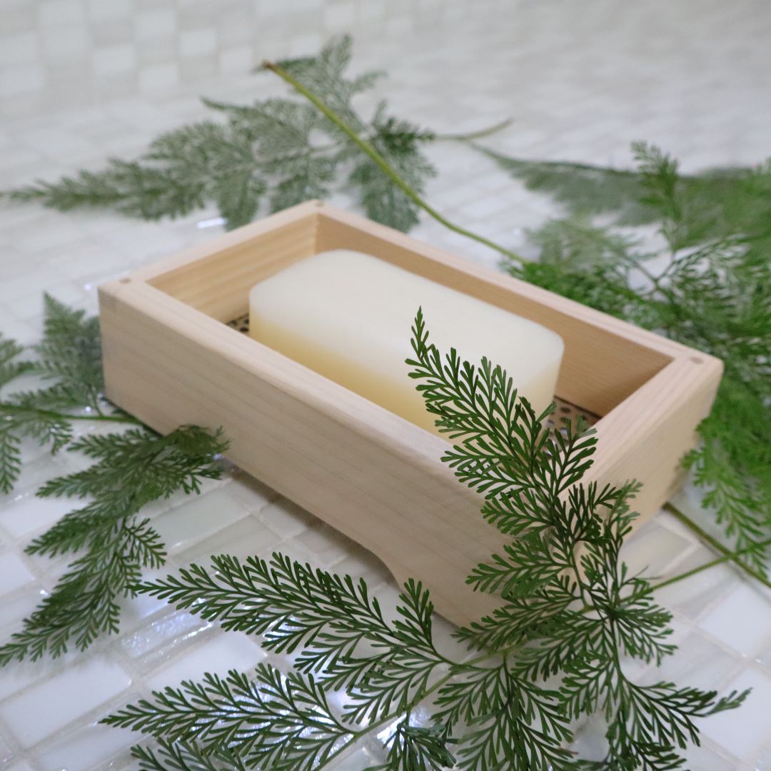  A pale wooden soap dish containing a bar of soap, nestled among fresh, green pine-like branches on a white tiled surface, evoking a natural and serene bathroom setting.