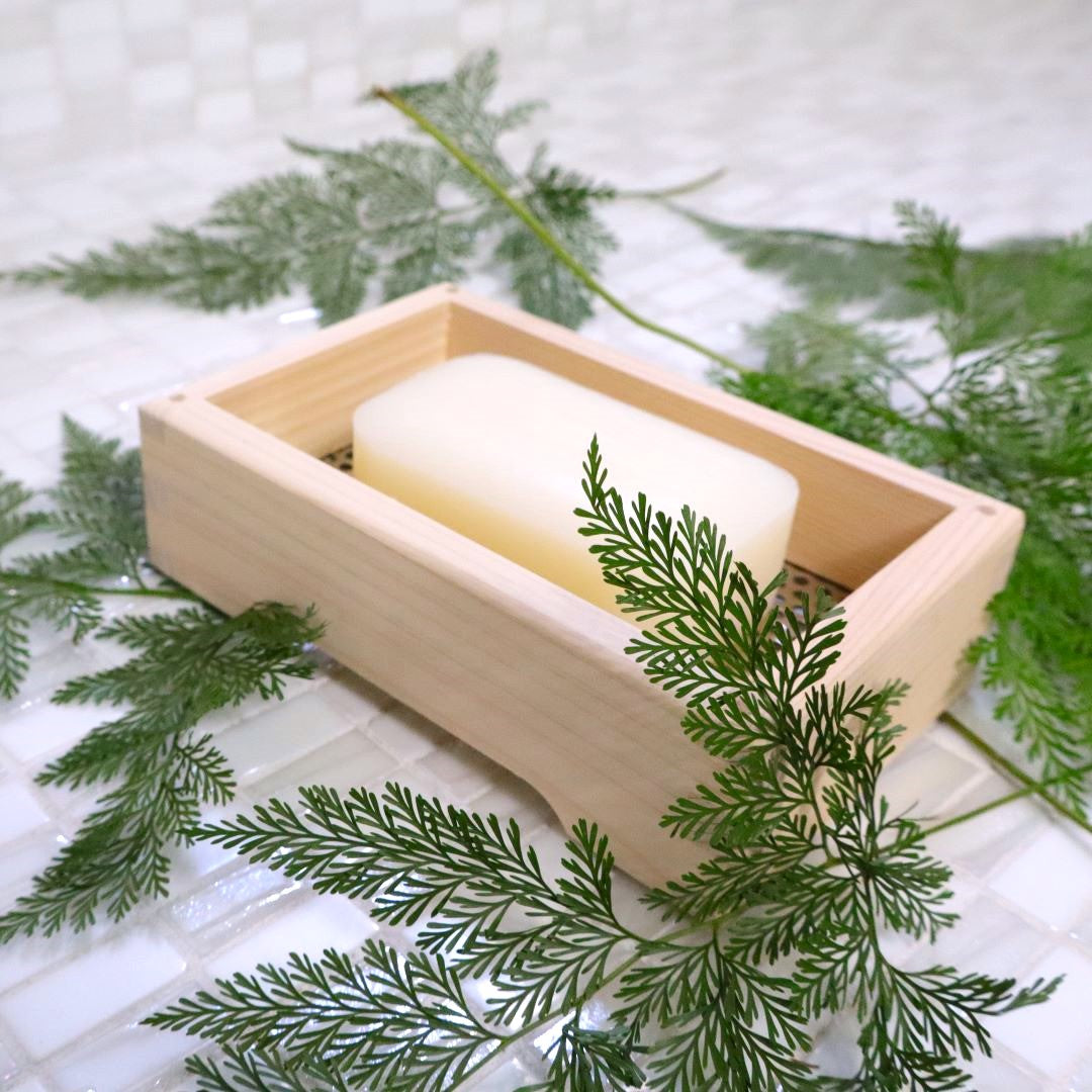 A wooden soap dish made of hinoki cypress, containing a pale, solid block of soap, placed on a tiled surface with a green pine branch arranged around it for an added natural touch.