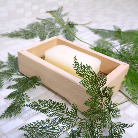  A wooden soap dish made of hinoki cypress, containing a pale, solid block of soap, placed on a tiled surface with a green pine branch arranged around it for an added natural touch.