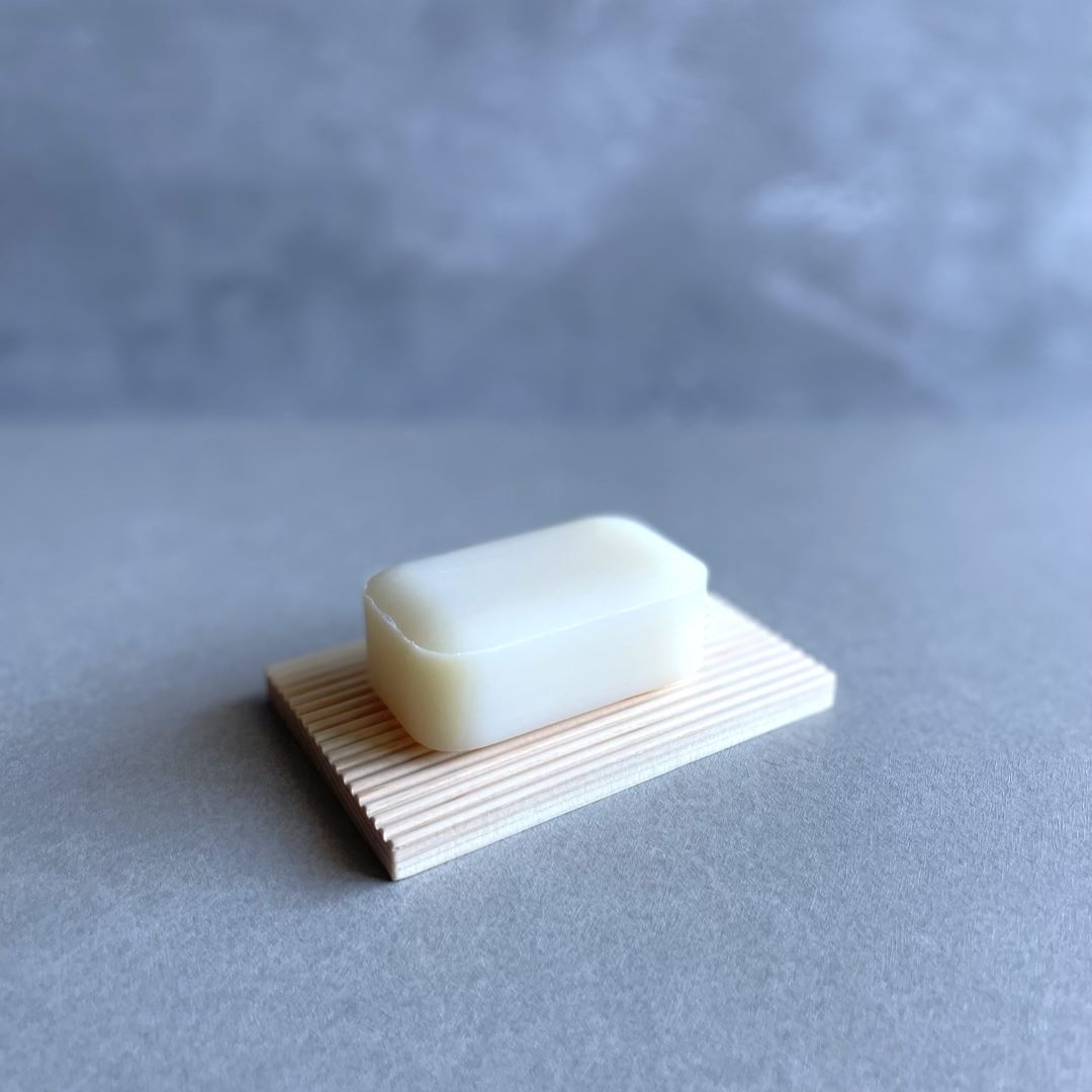  A bar of pale cream-colored soap rests on a light wooden soap dish with parallel ridges, positioned on a grey textured surface. The soap has a smooth, slightly worn surface, indicating it may have been used. The background is a uniform grey, providing a soft, neutral backdrop that highlights the simplicity of the soap and the natural design of the wooden soap dish.