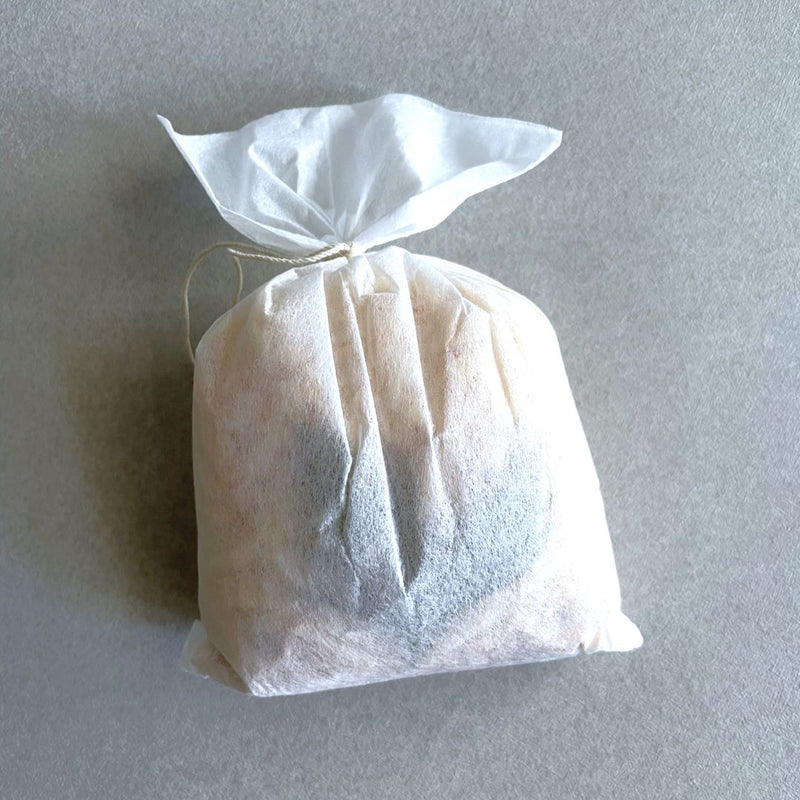 A white cloth bag tied with a thin string is placed on a textured grey background. The bag appears to contain a soft, possibly granular substance, creating rounded edges and a puffed appearance. The top of the bag is gathered and tied neatly, with the ends of the cloth and string extending upwards, giving it a hand-tied, artisanal look.