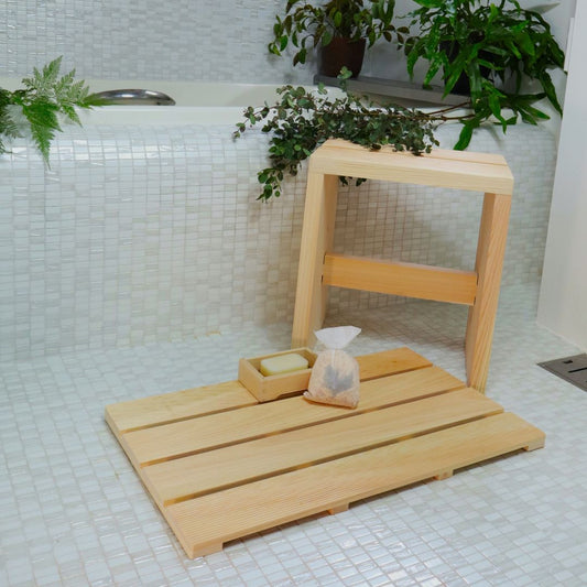 Eco-friendly bathroom accessories including a wooden stool, bath mat, soap dish with Hinoki soap, and spa aroma bags, surrounded by potted plants.