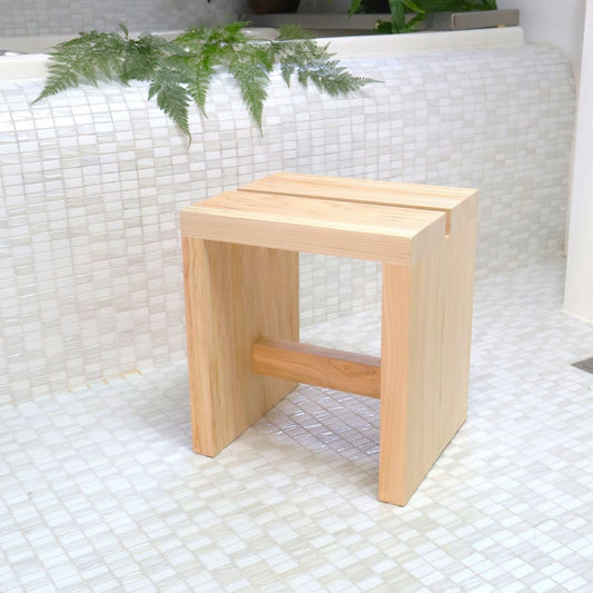A handcrafted Japanese cypress (Hinoki) bath chair, 11.8 inches tall, sits on a mosaic-tiled floor. Its eco-friendly, minimalist design is evident in the visible grain and natural finish, accompanied by subtle greenery in a serene bathroom setting.