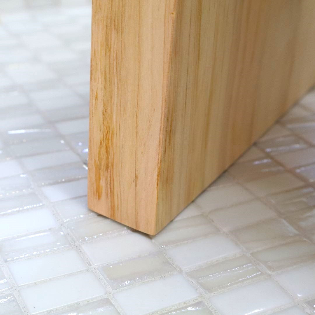 Close-up of a corner of a Hinoki wood bath chair on glossy white mosaic tiles, highlighting the chair&amp; natural wood grain and craftsmanship.