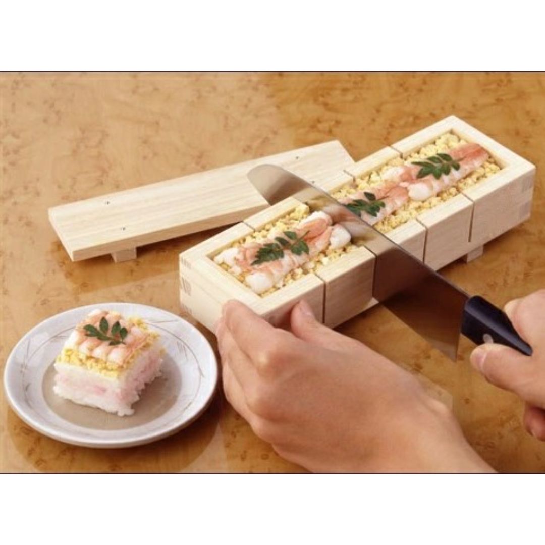 An hand hand holding a knife and cutting square sushi in a wooden sushi mold on a wooden table