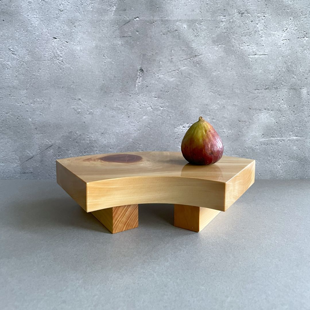 A single-tiered wooden sushi serving tray with sturdy legs, displaying a rich wood grain and featuring a single ripe fig on its surface, set against a grey backdrop with a concrete texture.
