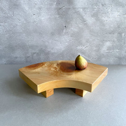 A single-tiered wooden sushi serving tray with sturdy legs, displaying a rich wood grain and featuring a single ripe fig on its surface, set against a grey backdrop with a concrete texture.
