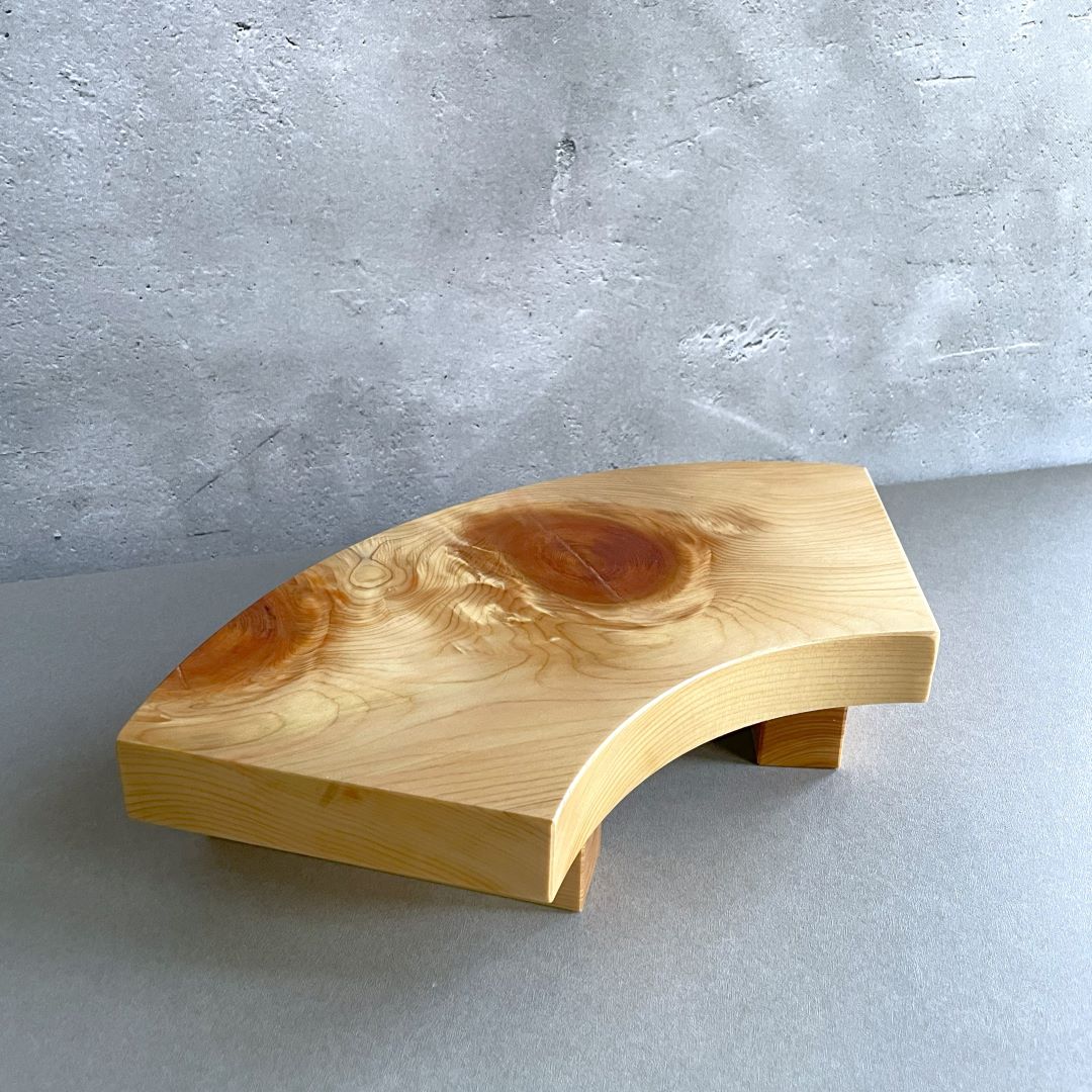  A wooden sushi serving tray with an elegant arch shape, featuring a prominent, beautifully patterned wood knot, resting on a grey surface against a textured wall.