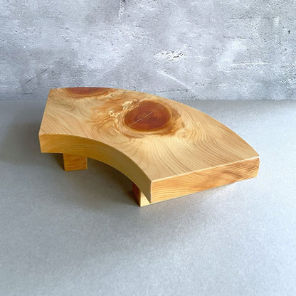 A curved wooden sushi serving tray showcasing a rich, swirling wood grain pattern, supported by a sturdy base, positioned on a grey surface with a textured concrete wall in the background.
