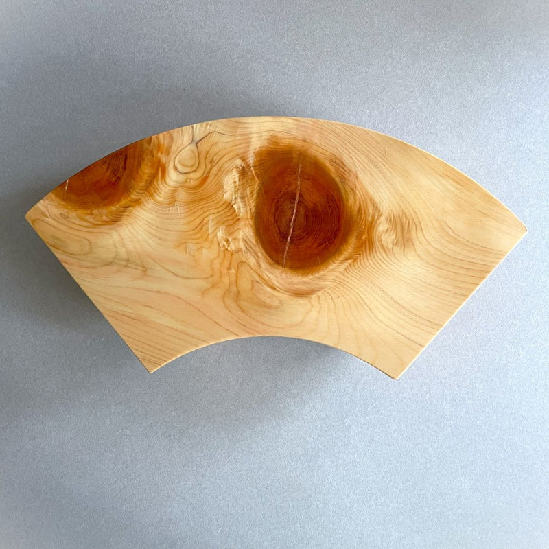 Close-up of a wooden sushi serving tray with a fan-like shape and a distinctive circular wood grain pattern, set against a grey surface and background.