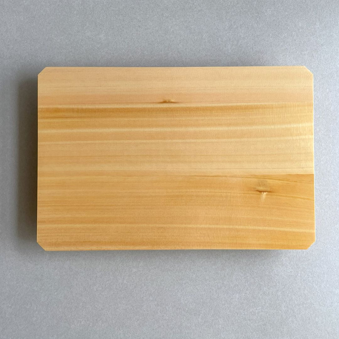 Top view of a rectangular Hinoki wood cutting board with a fine grain pattern, showcasing its natural light golden color. The board is placed on a grey countertop, highlighting the warm tones of the wood and its smooth, polished surface. The simple design of the board emphasizes its elegance and functional beauty.
