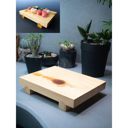 An overhead view of a wooden sushi tray placed on a gray kitchen counter. In the background, four small plants are visible, adding a touch of greenery to the scene. The backdrop consists of a plain gray wall, providing a simple and neutral background to the composition.