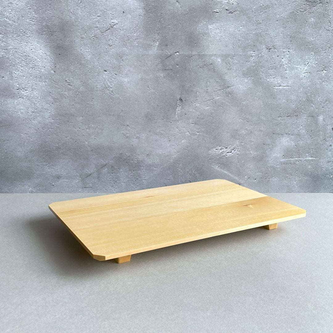 A simple, light-hued Hinoki wood sushi tray on a grey surface against a textured concrete wall. The tray is elevated slightly by its understated feet, showcasing its clean lines and smooth finish that exemplify minimalist design.