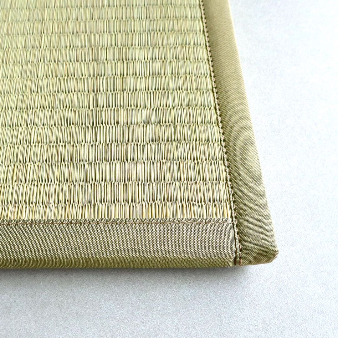 A close-up image of the corner of a tatami mat. The mat is made of tightly woven rush grass, with a reinforced edge made of fabric for durability. The stitching along the edge is visible, showcasing the craftsmanship of the mat. The background is a light, neutral color that highlights the texture and detail of the tatami mat.