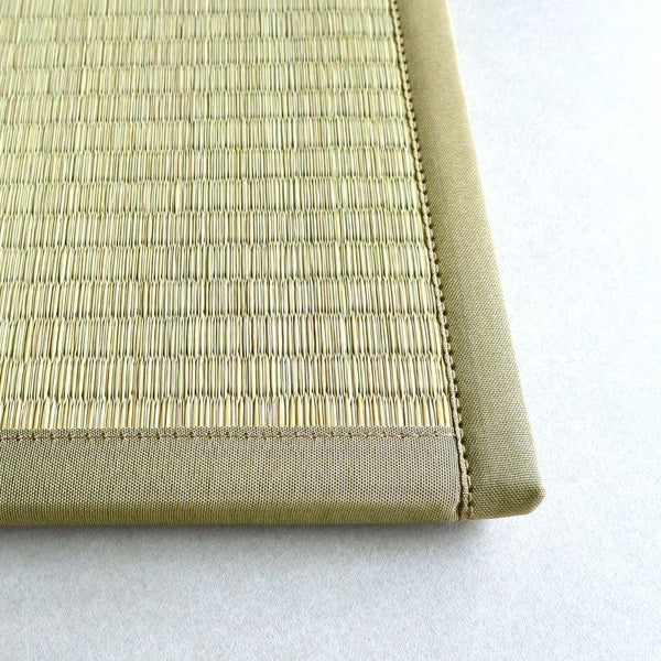 A close-up image of the corner of a tatami mat. The mat is made of tightly woven rush grass, with a reinforced edge made of fabric for durability. The stitching along the edge is visible, showcasing the craftsmanship of the mat. The background is a light, neutral color that highlights the texture and detail of the tatami mat.