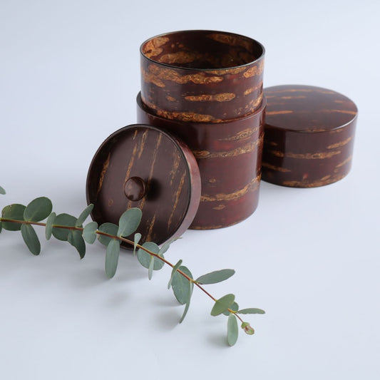 A natural wood tea or coffee caddy crafted from cherry bark, with a capacity of 200g. The container features a unique pattern with horizontal stripes, shown with an open lid beside it. A sprig of eucalyptus adds a touch of greenery.