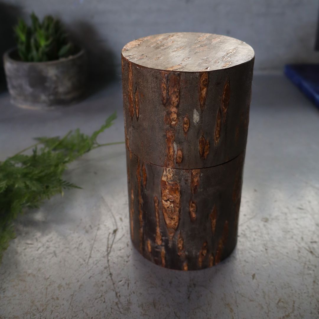 A cylindrical tea/coffee caddy made of natural wild cherry bark with a marbled exterior, displayed on a flat surface with a potted plant in the background.