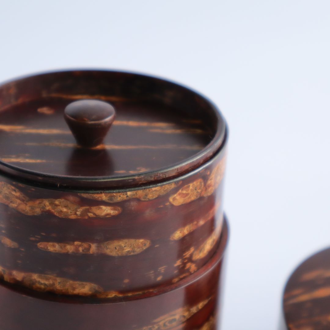 Close-up of the top part of a cherry bark tea caddy, showing the smooth, round knob on the lid and the horizontal stripes of the natural bark pattern. The focus is soft, with a gentle gradient to the white background.