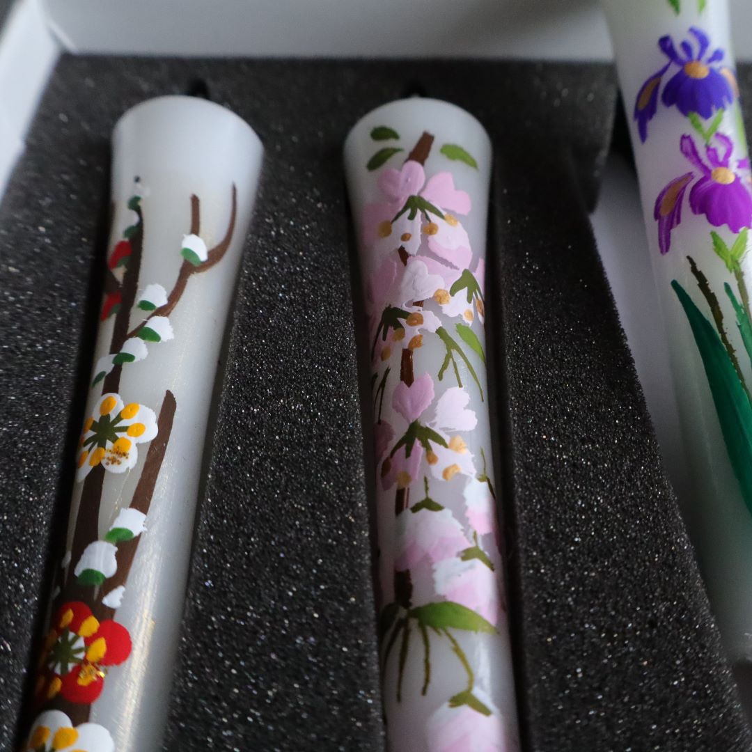 A close-up view of three hand-painted sumac wax candles with intricate floral designs, nestled securely in a box with black foam inserts. The visible candle designs feature a mix of daisies.