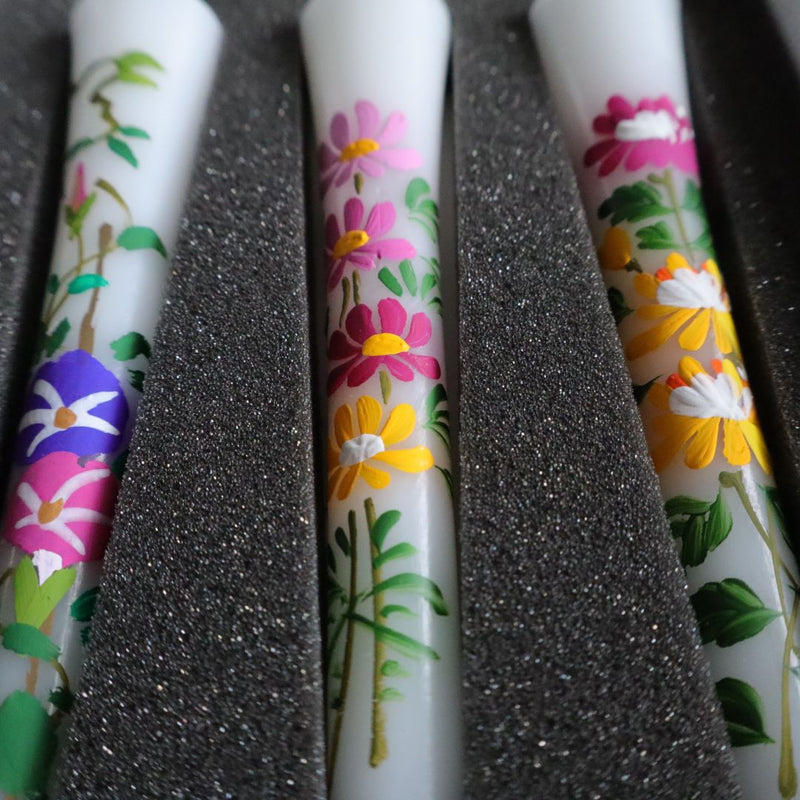 Close-up of three artisanal sumac wax candles with vibrant hand-painted floral designs, resting in a foam-lined box. The detailed flowers in hues of purple, pink, yellow, and white bloom against the candles&#39; white background.