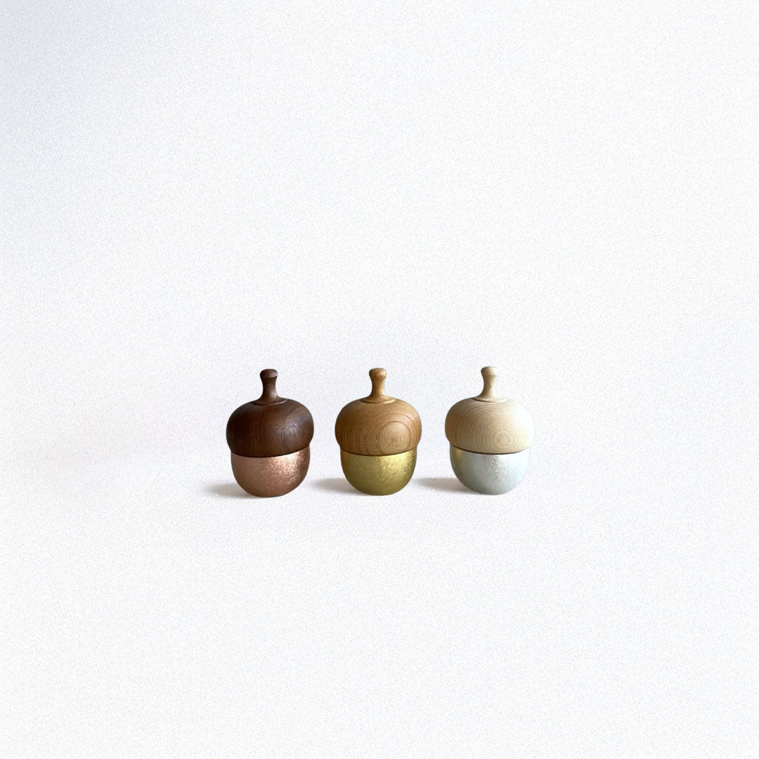 Three handcrafted Healing Bells standing in a row on a grey surface. The first one has a pink color, the second one is goldish, and the last one is white-grey. Each bell has an acorn shape with a wooden top and a brass pendulum tucked inside.