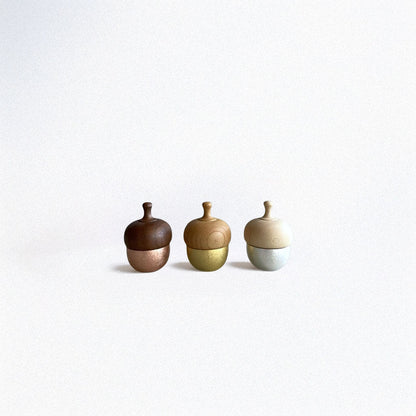Three handcrafted Healing Bells standing in a row on a grey surface. The first one has a pink color, the second one is goldish, and the last one is white-grey. Each bell has an acorn shape with a wooden top and a brass pendulum tucked inside.