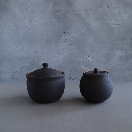 Two handmade black ceramic salt cellars with lids, one larger and one smaller, set against a textured grey background.