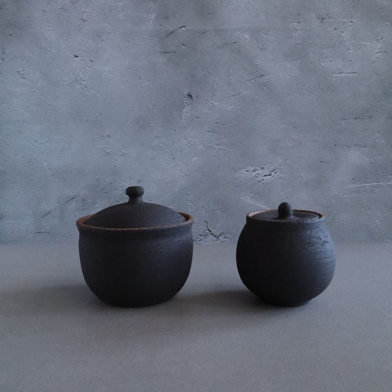 Two handmade black ceramic salt cellars with lids, one larger and one smaller, set against a textured grey background.