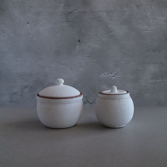 Two white handmade Japanese Shigaraki ceramic salt cellars with brown-trimmed lids on a gray background.