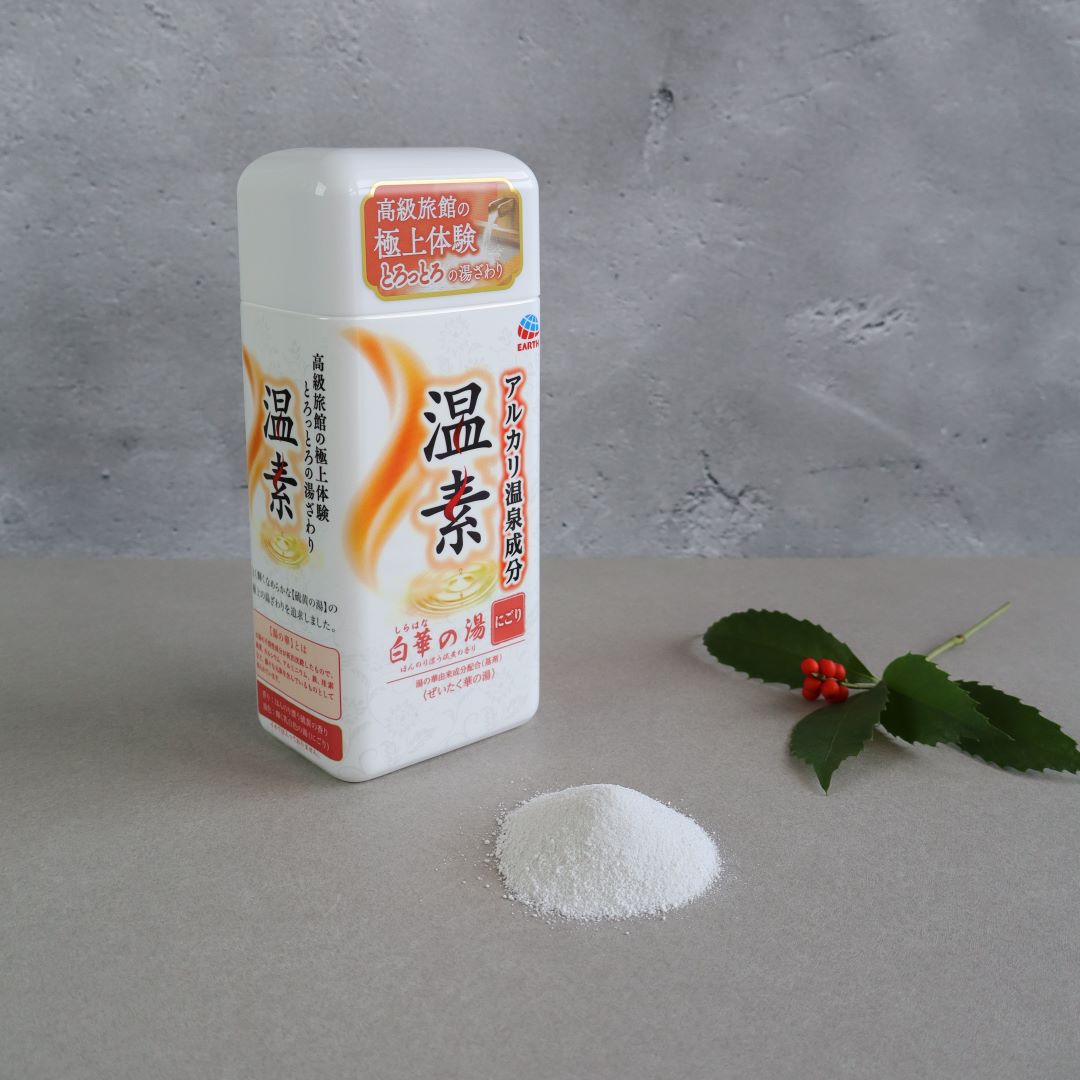 A container of Onso Shiraha no Yu Bath Salts with a white and orange design, placed on a neutral surface. A small pile of fine white bath powder is beside it, along with a sprig of green leaves and red berries, creating a natural and calming aesthetic.