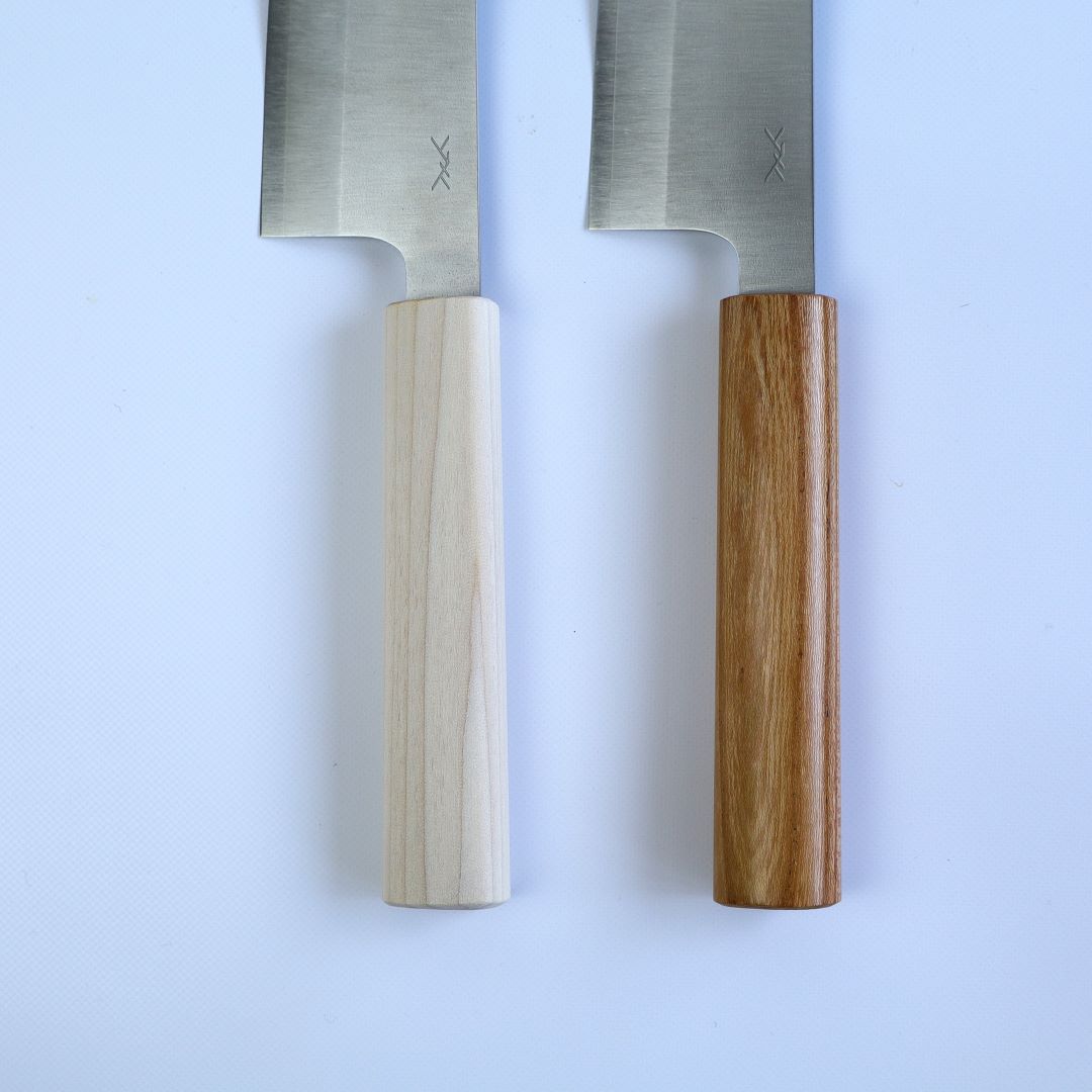 Two Santoku knives with the blades pointing upwards, showcasing their wooden handles against a white background. The knife on the left features a pale, light wood handle, while the knife on the right has a darker, richly grained wood handle. Both handles are cylindrical, with a visible grain pattern, complementing the clean lines of the brushed stainless steel blades which bear a simple, elegant maker's mark.