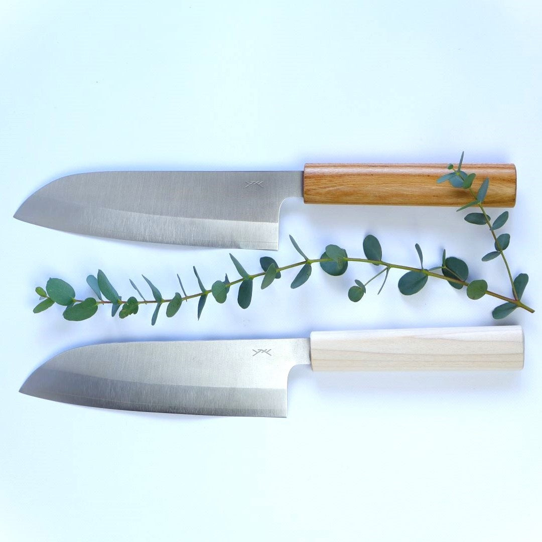  Two premium Japanese Santoku knives with wooden handles, lying flat on a white background, accented by a sprig of green eucalyptus leaves. The top knife has a rich, honey-toned handle, while the bottom knife features a pale, natural wood handle. Both knives have a smooth, matte-finish stainless steel blade with a visible sharp edge, and the manufacturer's mark subtly etched near the handle.