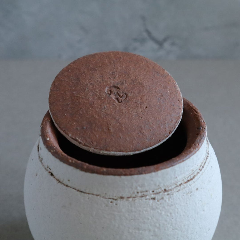 Close-up of a white Shigaraki ceramic container opening with a round, unglazed terra cotta lid partially covering it. The lid has a stamped symbol, and the rim of the container has a rustic brown accent, set against a gray concrete background.