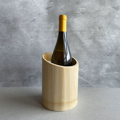 A bottle of wine with a yellow foil capsule is standing in a cylindrical, light wooden wine cooler. The cooler and bottle are placed on a gray surface with a textured gray concrete wall in the background.