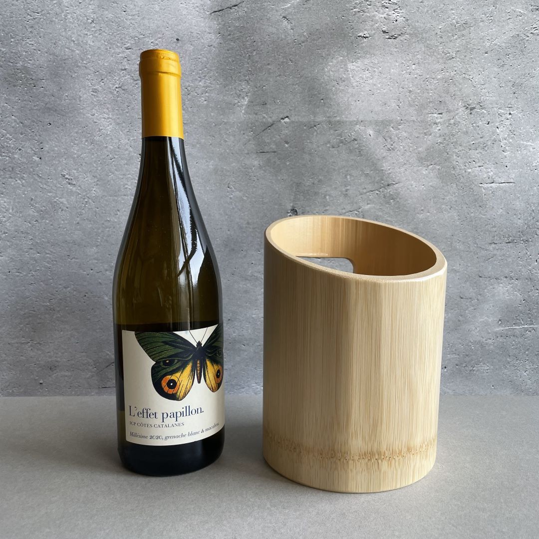 A wine bottle with a yellow cap and a label featuring a butterfly illustration is standing next to a cylindrical, light wooden wine cooler. They are placed on a gray surface with a textured gray concrete wall in the background. The label on the bottle reads "L'effet papillon," indicating a French origin.