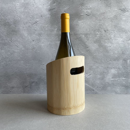 A wine bottle with a yellow cap is partially encased in a modern, light wooden wine cooler with a handle cutout. The wine cooler is on a gray surface against a textured concrete wall background.
