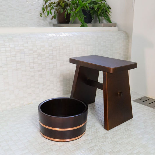 A large black wooden shower stool paired with a matching Hinoki Cypress bucket, both with copper accents, on a tiled bathroom floor.